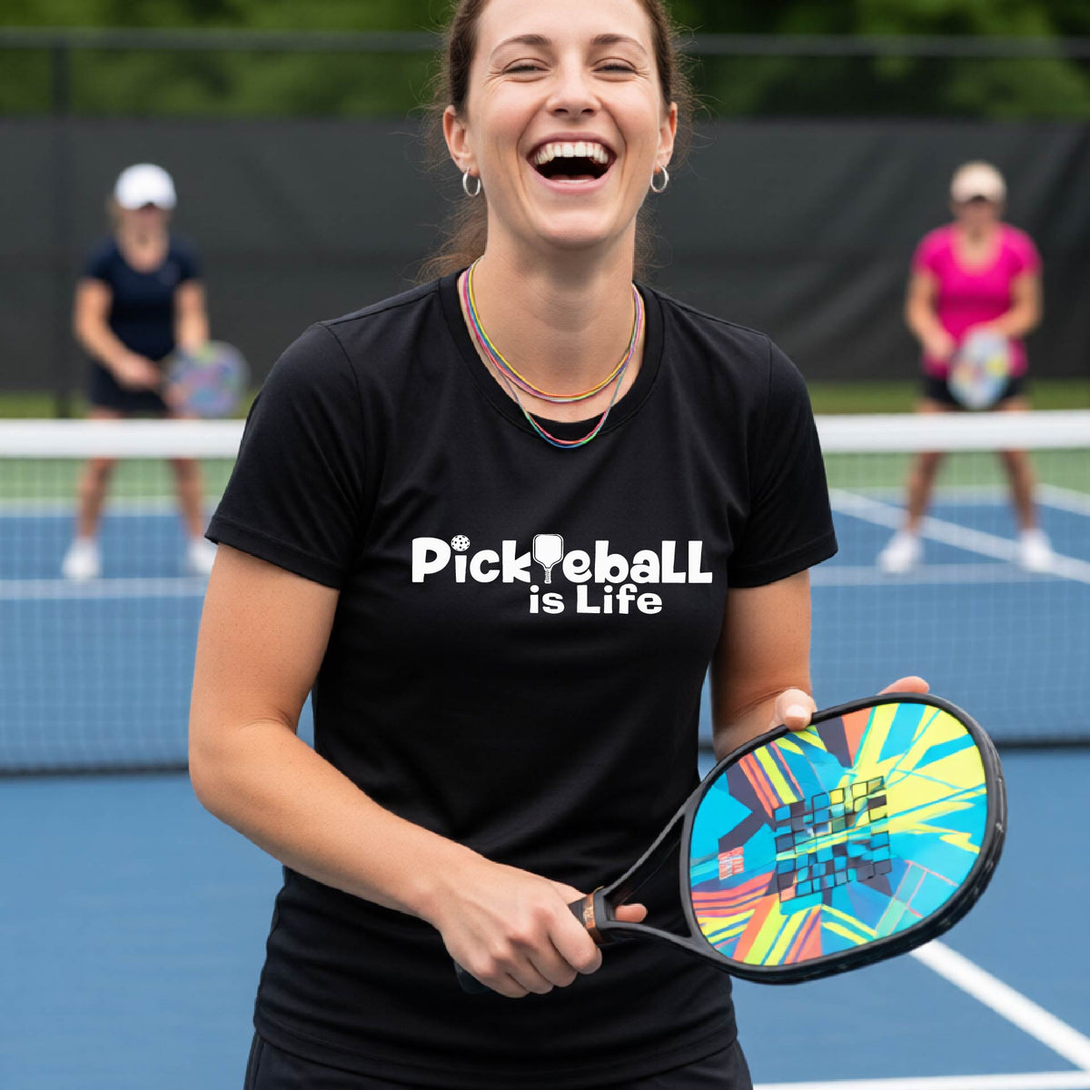 A woman on a pickleball court wearing a black short-sleeve t-shirt  with the text 'Pickleball is Life' with the L replaced with a pickleball paddle.