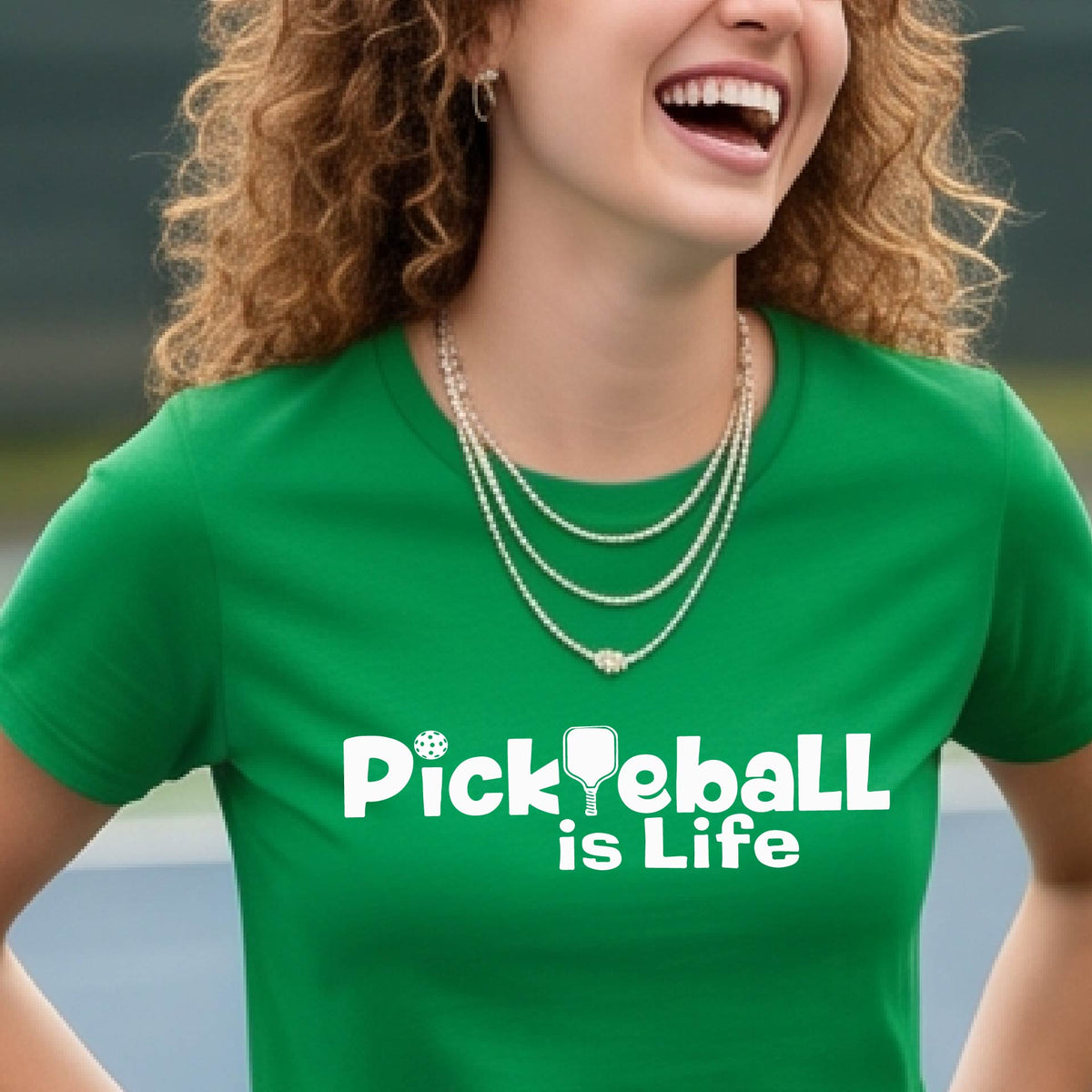 A woman on a pickleball court wearing a green short-sleeve t-shirt  with the text 'Pickleball is Life' with the L replaced with a pickleball paddle.