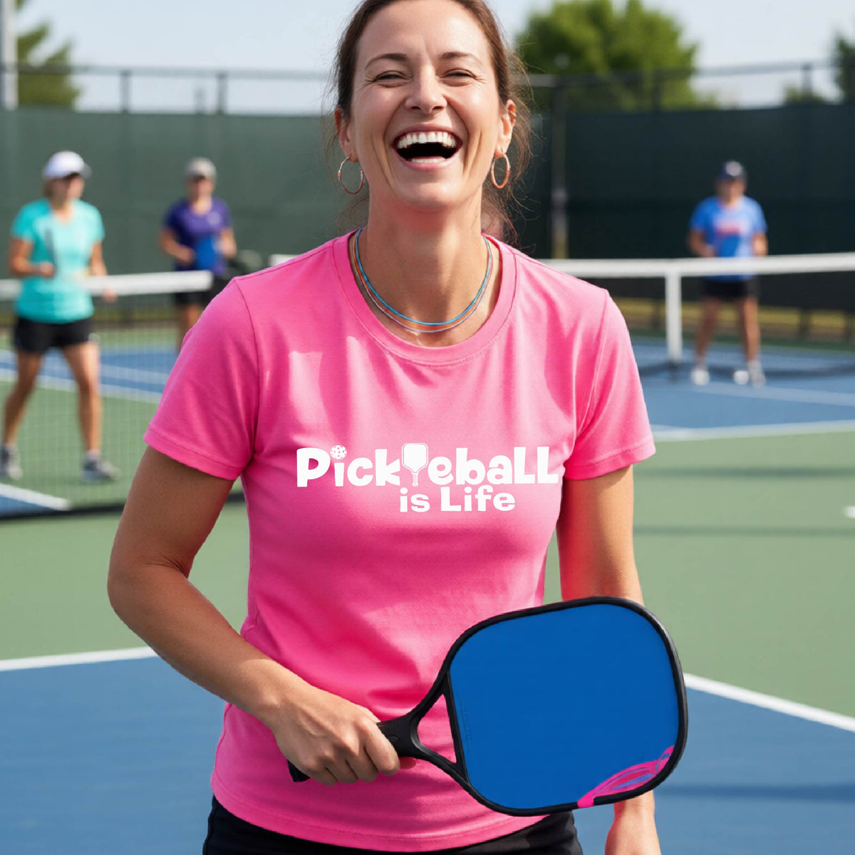 A woman on a pickleball court wearing a bright pink short-sleeve t-shirt  with the text 'Pickleball is Life' with the L replaced with a pickleball paddle.