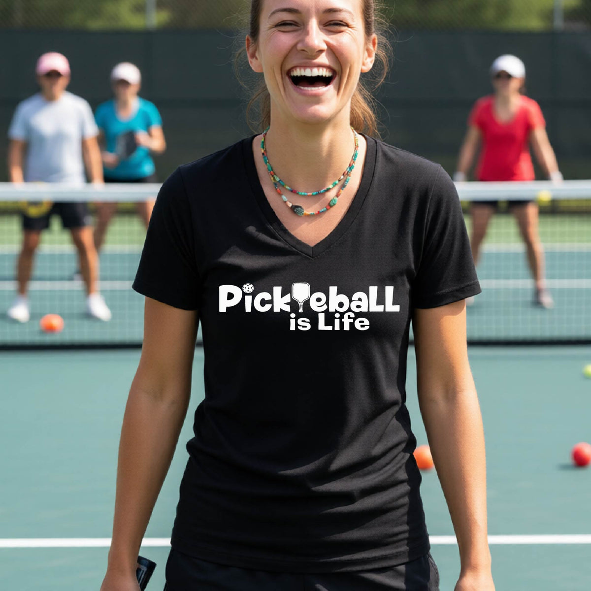 A woman on a pickleball court wearing a black short-sleeve v-neck t-shirt with the text 'Pickleball is Life' with the L replaced with a pickleball paddle.