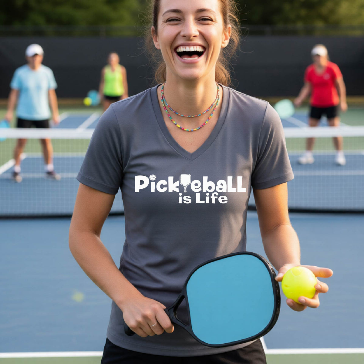 A woman on a pickleball court wearing a gray short-sleeve v-neck t-shirt with the text 'Pickleball is Life' with the L replaced with a pickleball paddle.