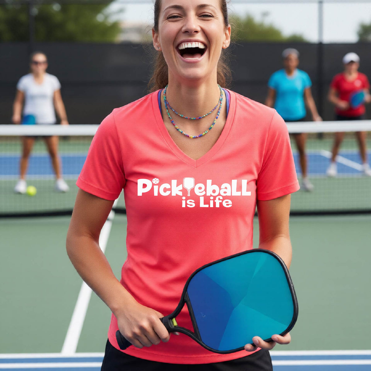 A woman on a pickleball court wearing a coral pink short-sleeve v-neck t-shirt with the text 'Pickleball is Life' with the L replaced with a pickleball paddle.