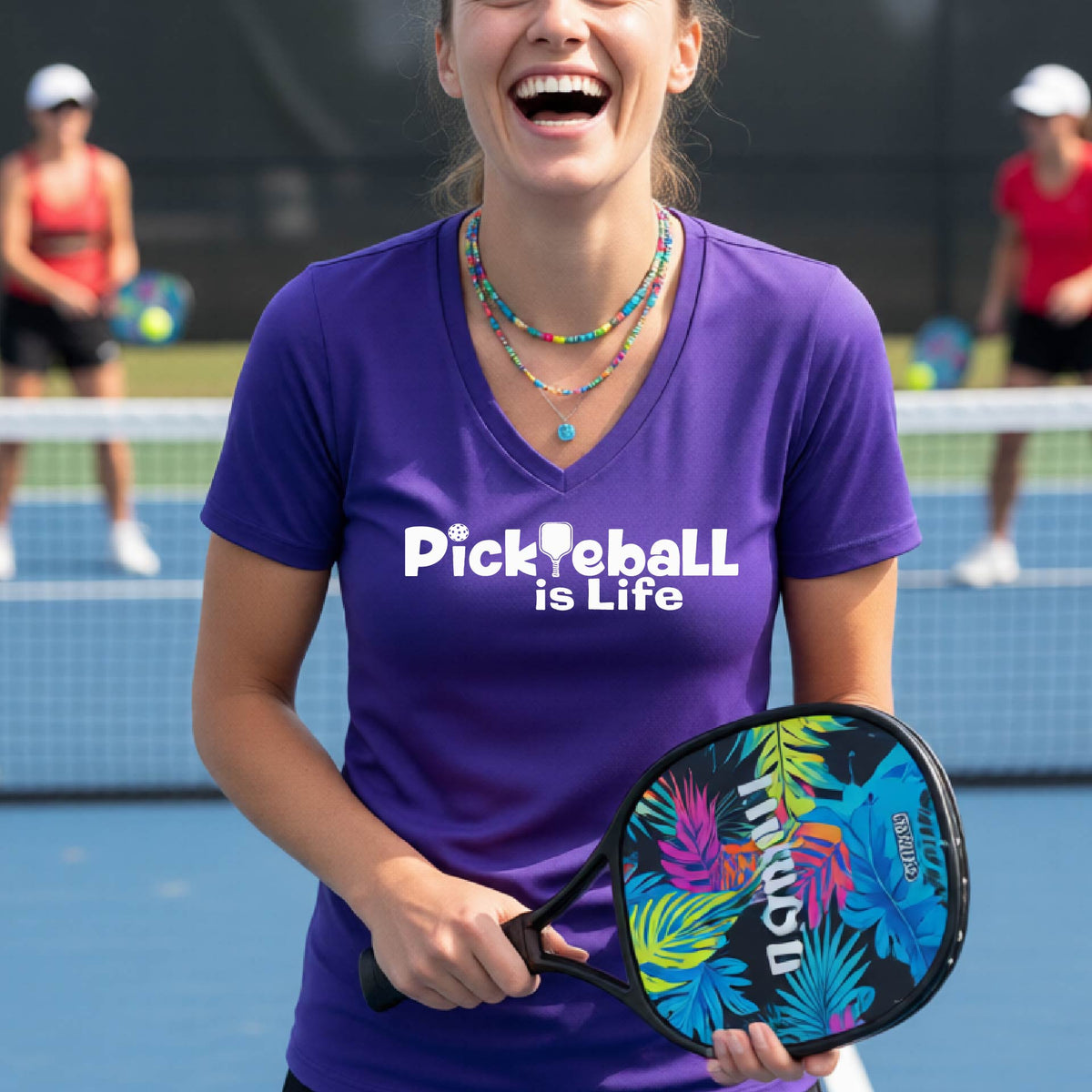 A woman on a pickleball court wearing a purple short-sleeve v-neck t-shirt with the text 'Pickleball is Life' with the L replaced with a pickleball paddle.