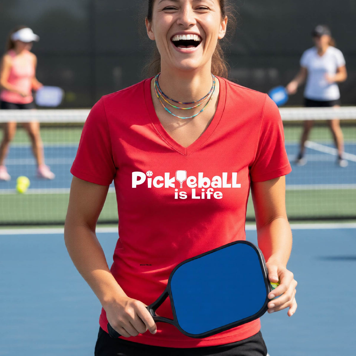 A woman on a pickleball court wearing a red short-sleeve v-neck t-shirt with the text 'Pickleball is Life' with the L replaced with a pickleball paddle.
