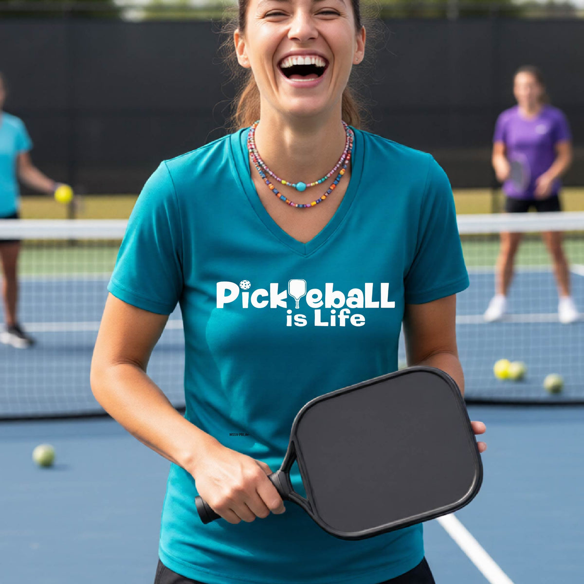 A woman on a pickleball court wearing a teal short-sleeve v-neck t-shirt with the text 'Pickleball is Life' with the L replaced with a pickleball paddle.