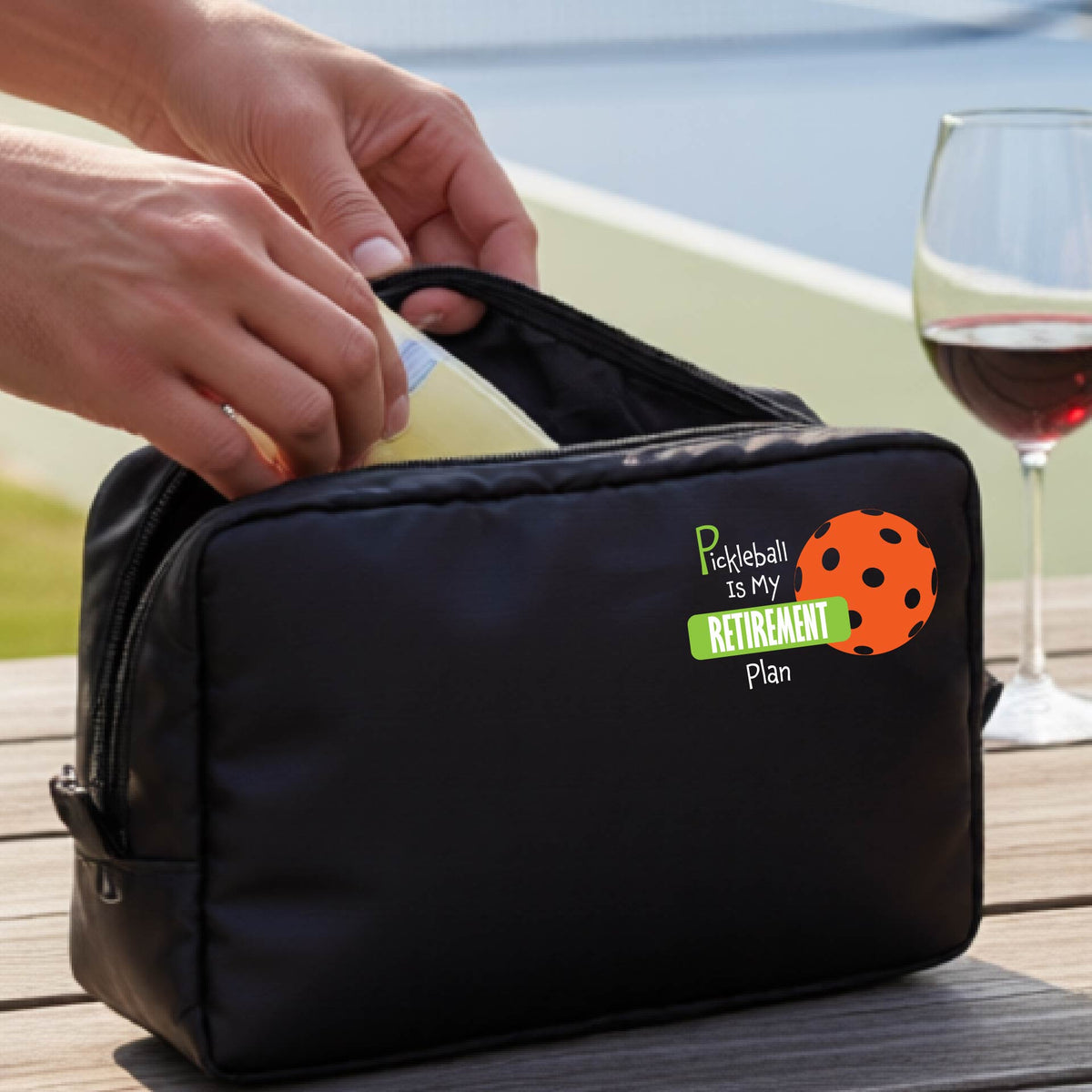 A close up of a black large cosmetic toiletry bag on a pickleball court, with a graphic of the words “Pickleball is my Retirement Plan” in green and white with an orange pickleball illustration in the background.