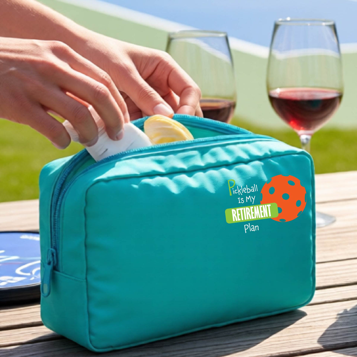 A close up of a turquoise large cosmetic toiletry bag on a pickleball court, with a graphic of the words “Pickleball is my Retirement Plan” in green and white with an orange pickleball illustration in the background.