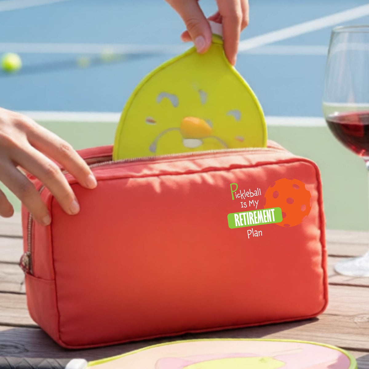 A close up of a coral colored large cosmetic toiletry bag on a pickleball court, with a graphic of the words “Pickleball is my Retirement Plan” in green and white with an orange pickleball illustration in the background.