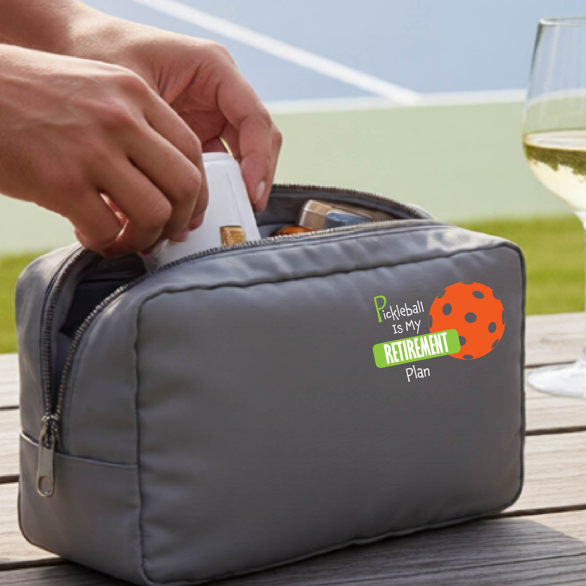 A close up of a gray large cosmetic toiletry bag on a pickleball court, with a graphic of the words “Pickleball is my Retirement Plan” in green and white with an orange pickleball illustration in the background.