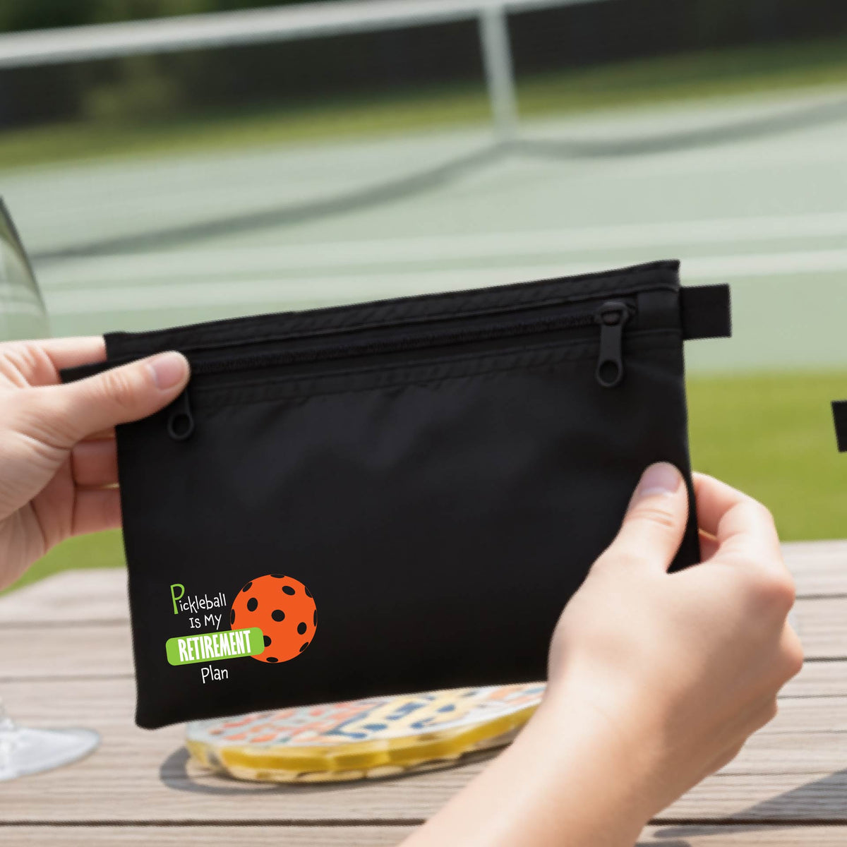 A close up of a black small cosmetic toiletry bag on a pickleball court, with a graphic of the words “Pickleball is my Retirement Plan” in green and white with an orange pickleball illustration in the background.