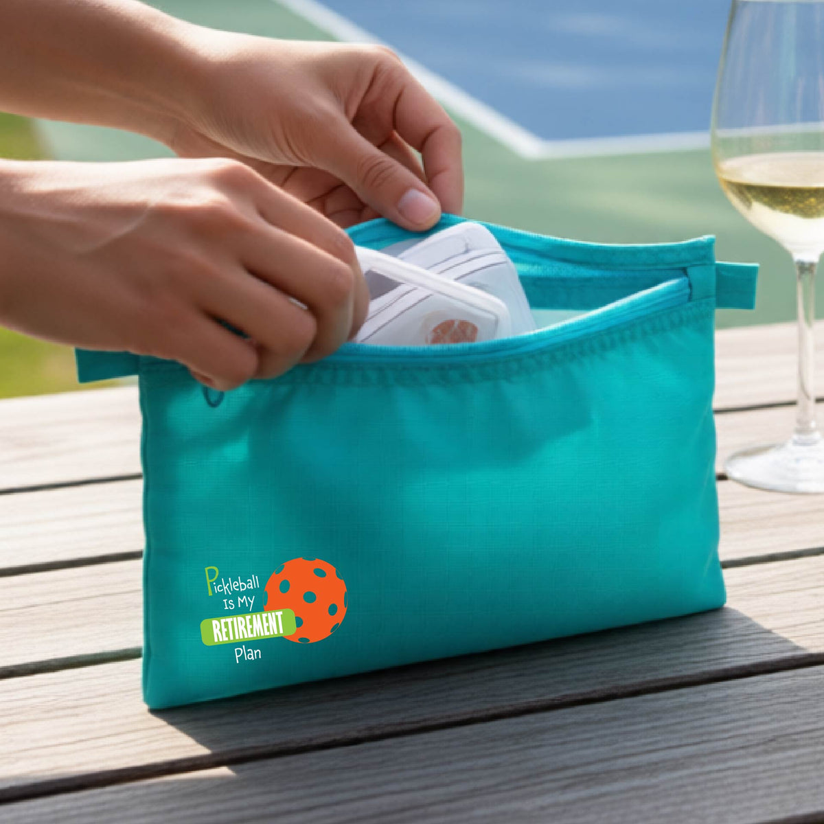 A close up of a turquoise small cosmetic toiletry bag on a pickleball court, with a graphic of the words “Pickleball is my Retirement Plan” in green and white with an orange pickleball illustration in the background.