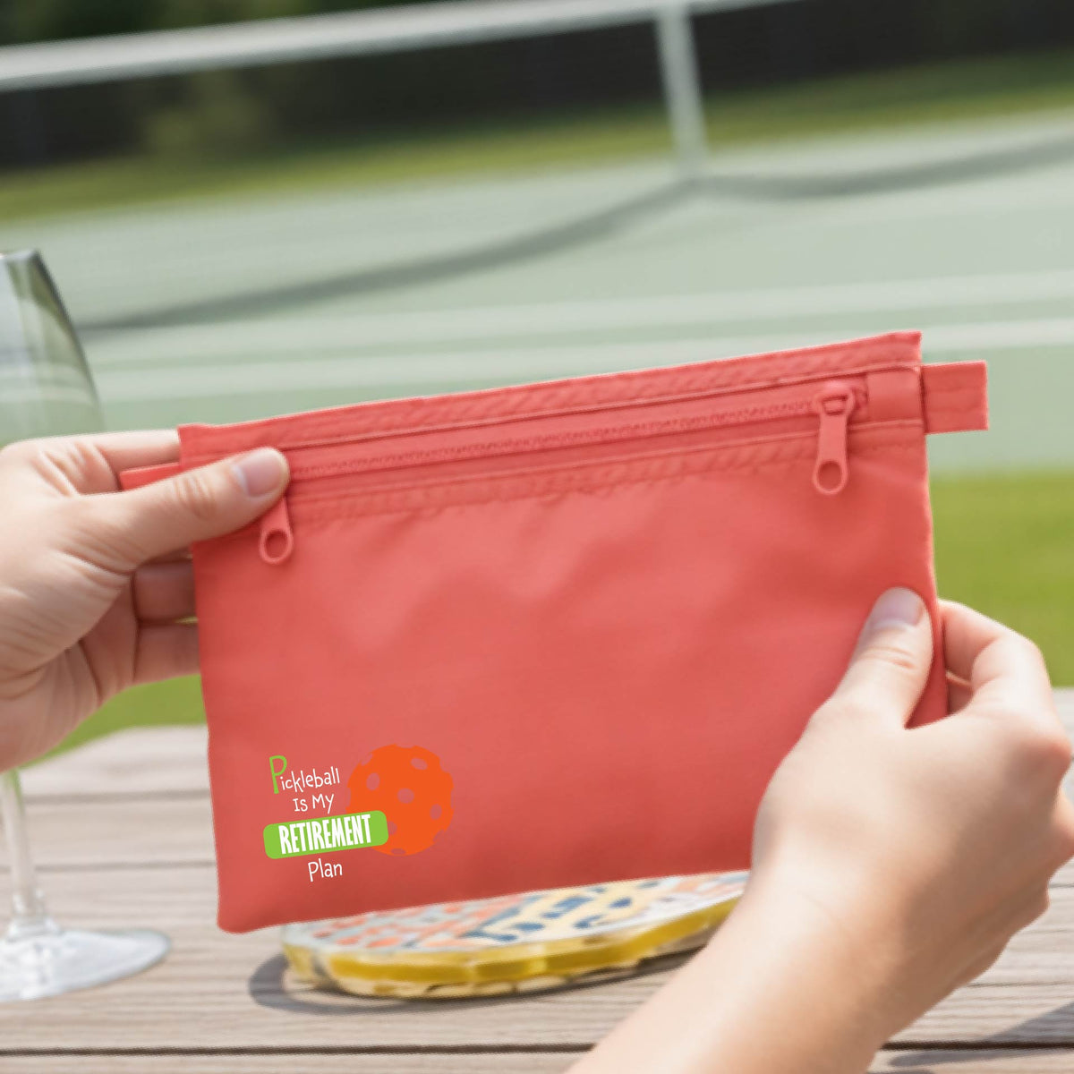 A close up of a coral colored small cosmetic toiletry bag on a pickleball court, with a graphic of the words “Pickleball is my Retirement Plan” in green and white with an orange pickleball illustration in the background.