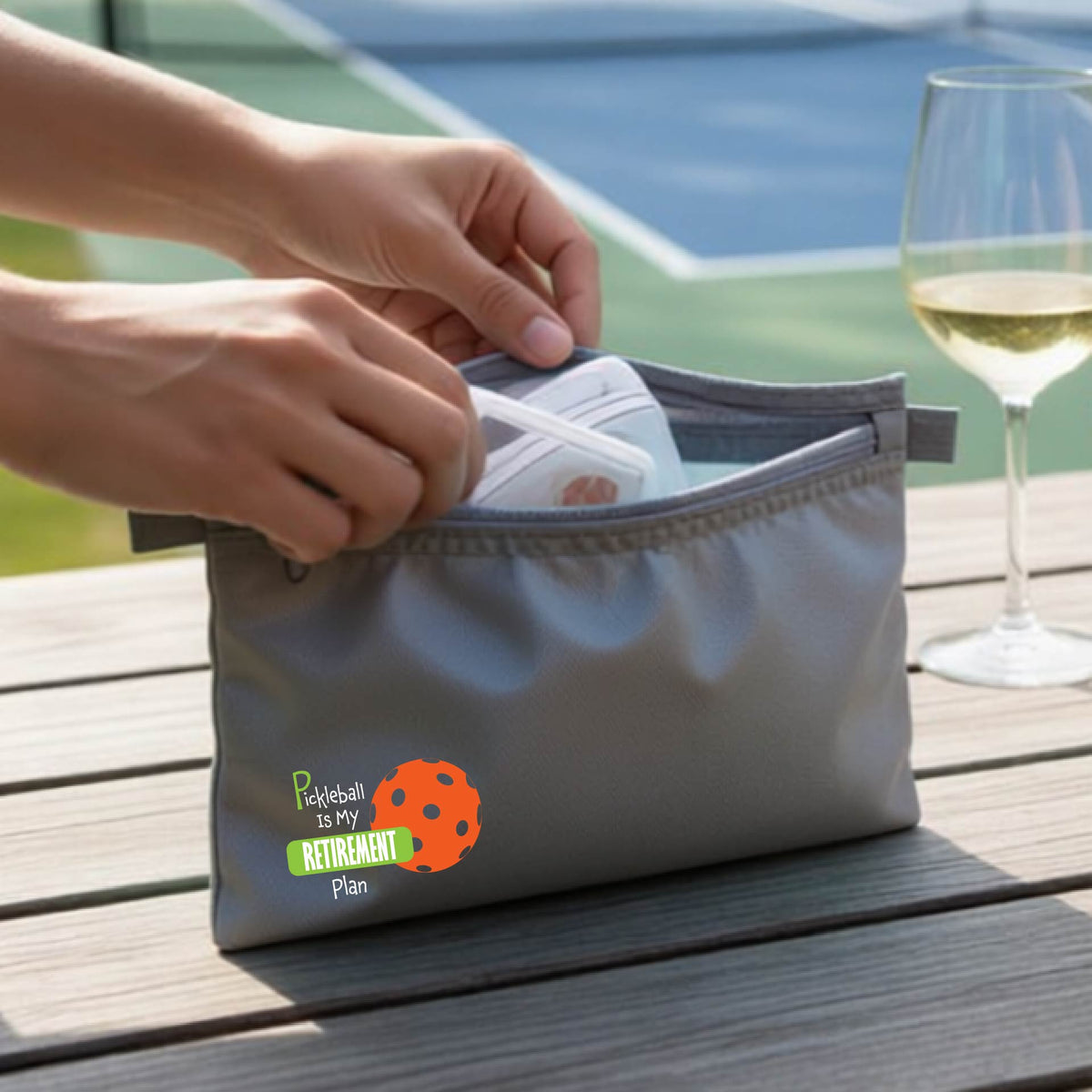 A close up of a gray small cosmetic toiletry bag on a pickleball court, with a graphic of the words “Pickleball is my Retirement Plan” in green and white with an orange pickleball illustration in the background.