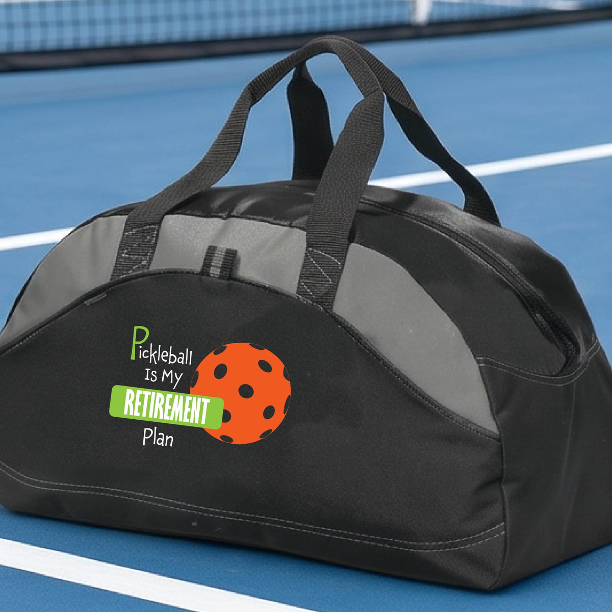 A close-up of a black duffel bag with gray panels. on a pickleball court, with a graphic of the words “Pickleball is my Retirement Plan” in green and white with an orange pickleball illustration in the background.