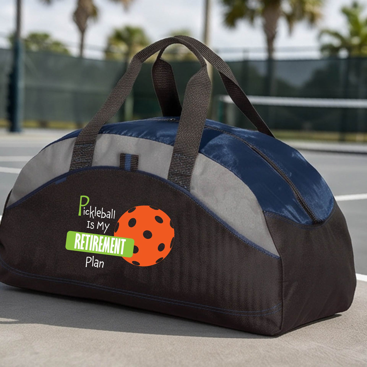 A close-up of a navy blue and black duffel bag with gray panels. on a pickleball court, with a graphic of the words “Pickleball is my Retirement Plan” in green and white with an orange pickleball illustration in the background.