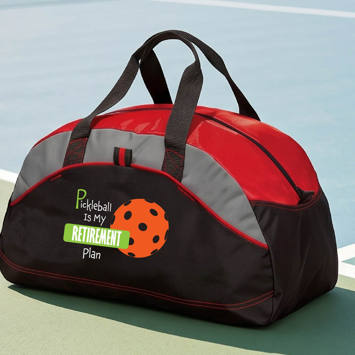 A close-up of a red and black duffel bag with gray panels. on a pickleball court, with a graphic of the words “Pickleball is my Retirement Plan” in green and white with an orange pickleball illustration in the background.