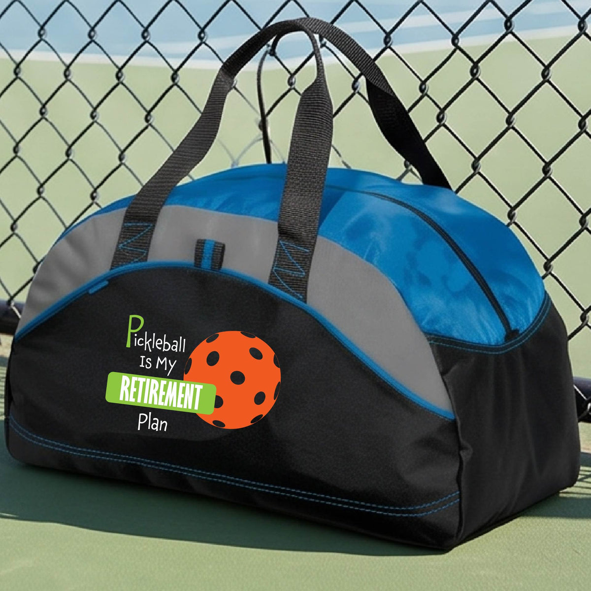 A close-up of a royal blue and black duffel bag with gray panels. on a pickleball court, with a graphic of the words “Pickleball is my Retirement Plan” in green and white with an orange pickleball illustration in the background.