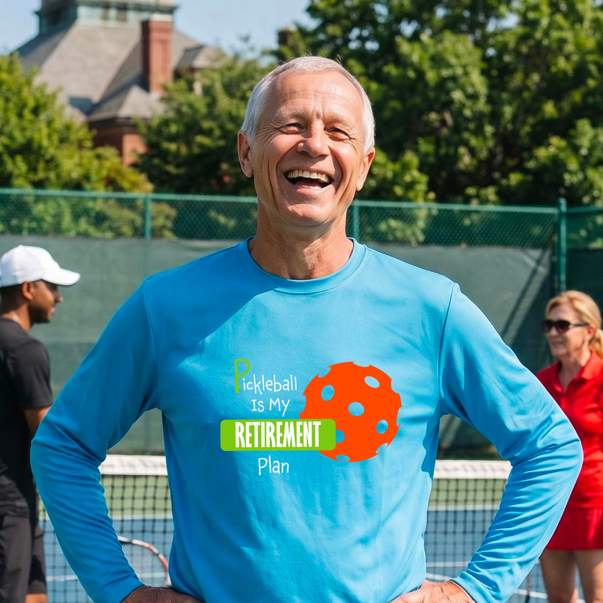 A smiling man stands on a pickleball court wearing a bright blue long-sleeve t-shirt with a graphic that reads 'Pickleball is my Retirement Plan' alongside an orange pickleball illustration. 
