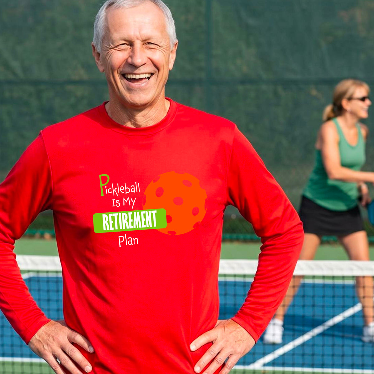 A smiling man stands on a pickleball court wearing a red long-sleeve t-shirt with a graphic that reads 'Pickleball is my Retirement Plan' alongside an orange pickleball illustration. 