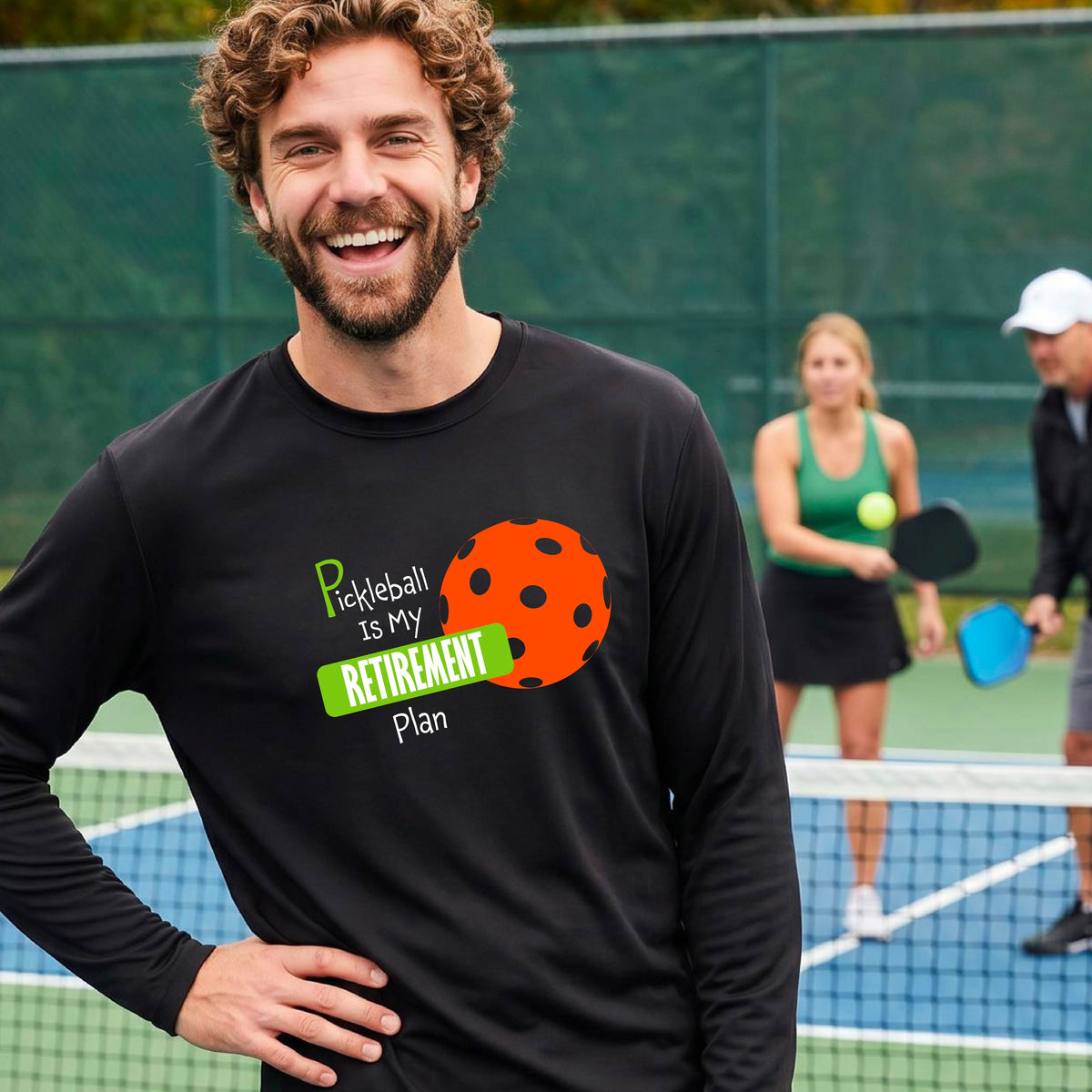 A smiling man stands on a pickleball court wearing a black long-sleeve t-shirt with a graphic that reads 'Pickleball is my Retirement Plan' alongside an orange pickleball illustration. 