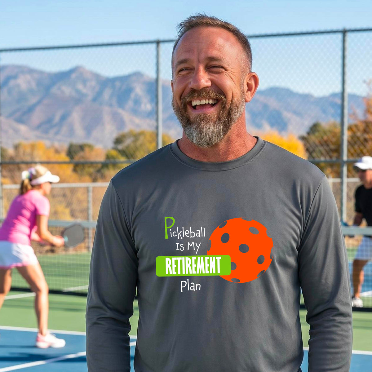 A smiling man stands on a pickleball court wearing a dark gray long-sleeve t-shirt with a graphic that reads 'Pickleball is my Retirement Plan' alongside an orange pickleball illustration. 