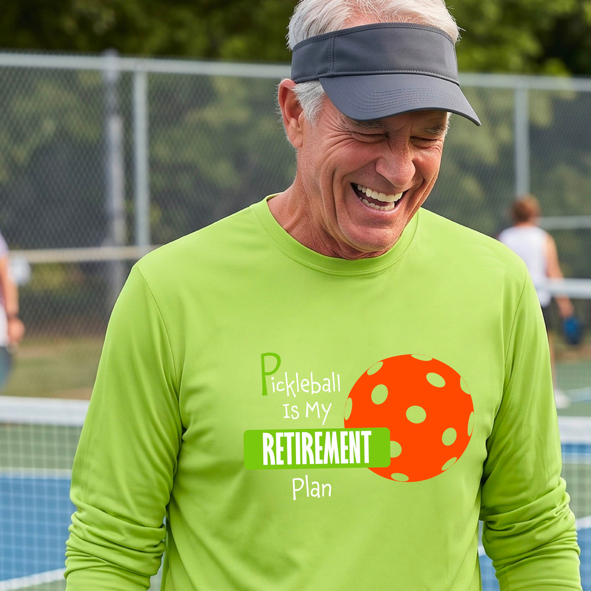 A smiling man stands on a pickleball court wearing a lime green long-sleeve t-shirt with a graphic that reads 'Pickleball is my Retirement Plan' alongside an orange pickleball illustration. 