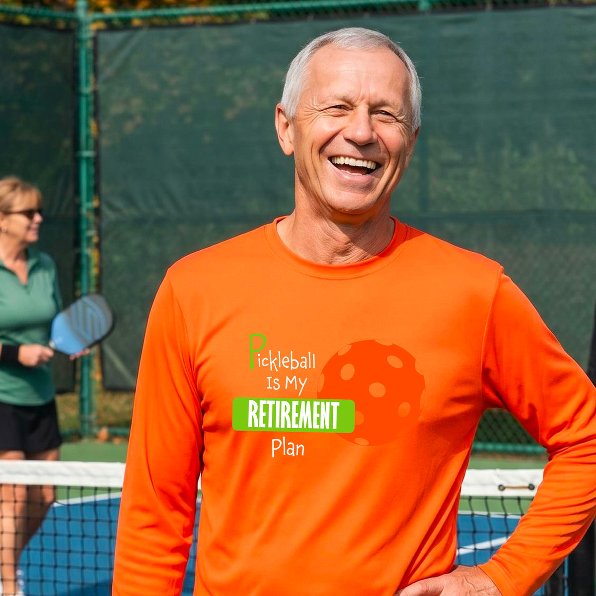 A smiling man stands on a pickleball court wearing a bright orange long-sleeve t-shirt with a graphic that reads 'Pickleball is my Retirement Plan' alongside an orange pickleball illustration. 