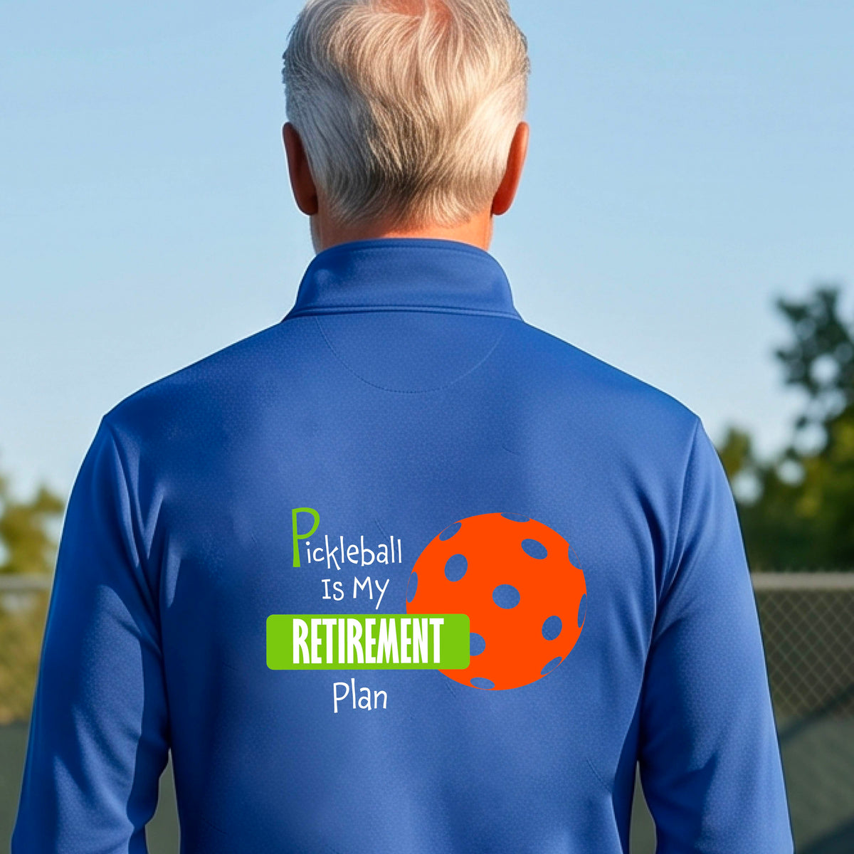 A man on a pickleball court, showing the back of a royal blue quarter-zip shirt with the words “Pickleball is my Retirement Plan” in green and white with an orange pickleball illustration in the background