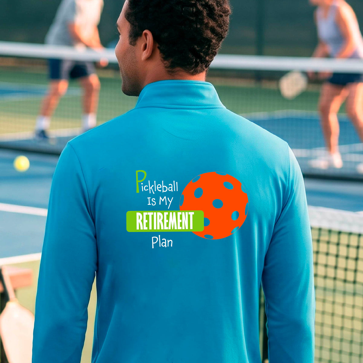 A man on a pickleball court, showing the back of a bright blue quarter-zip shirt with the words “Pickleball is my Retirement Plan” in green and white with an orange pickleball illustration in the background