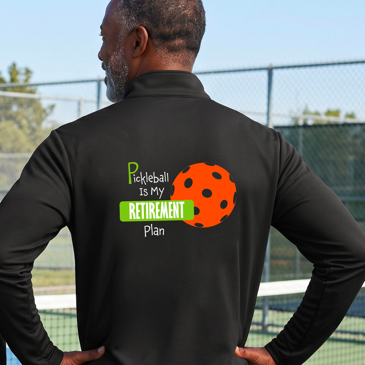A man on a pickleball court, showing the back of a black quarter-zip shirt with the words “Pickleball is my Retirement Plan” in green and white with an orange pickleball illustration in the background