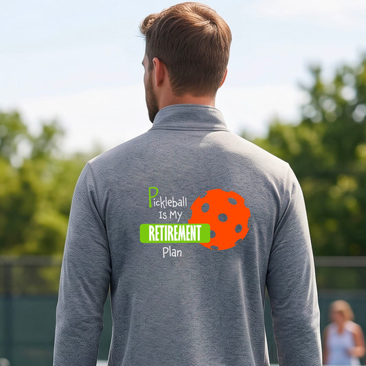 A man on a pickleball court, showing the back of a qray quarter-zip shirt with the words “Pickleball is my Retirement Plan” in green and white with an orange pickleball illustration in the background