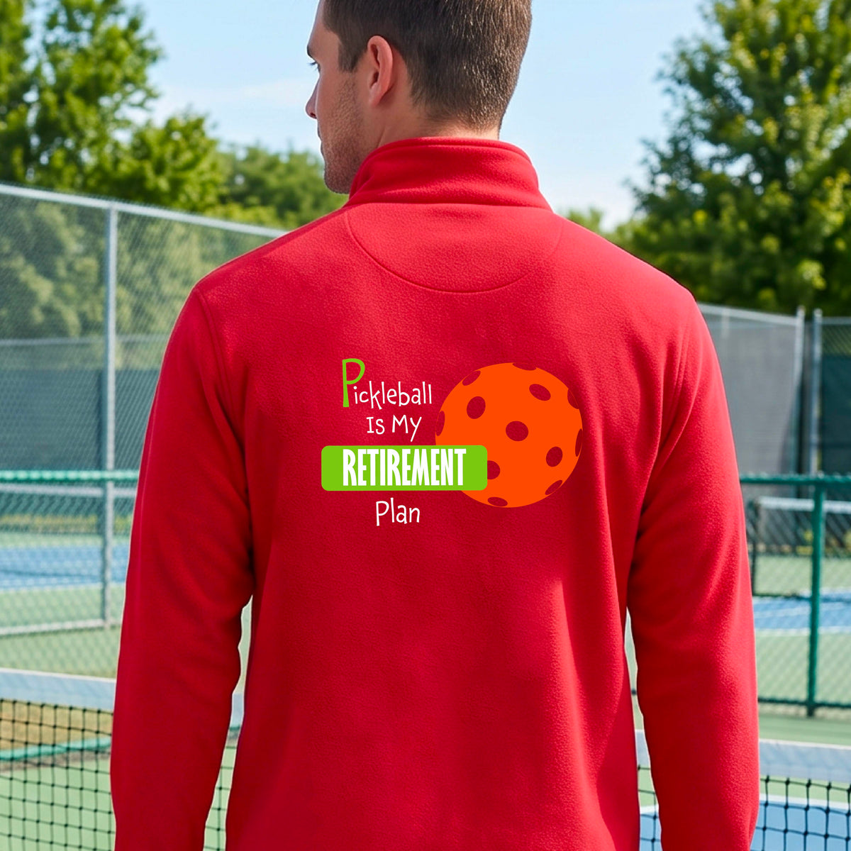 A man on a pickleball court, showing the back of a red quarter-zip shirt with the words “Pickleball is my Retirement Plan” in green and white with an orange pickleball illustration in the background