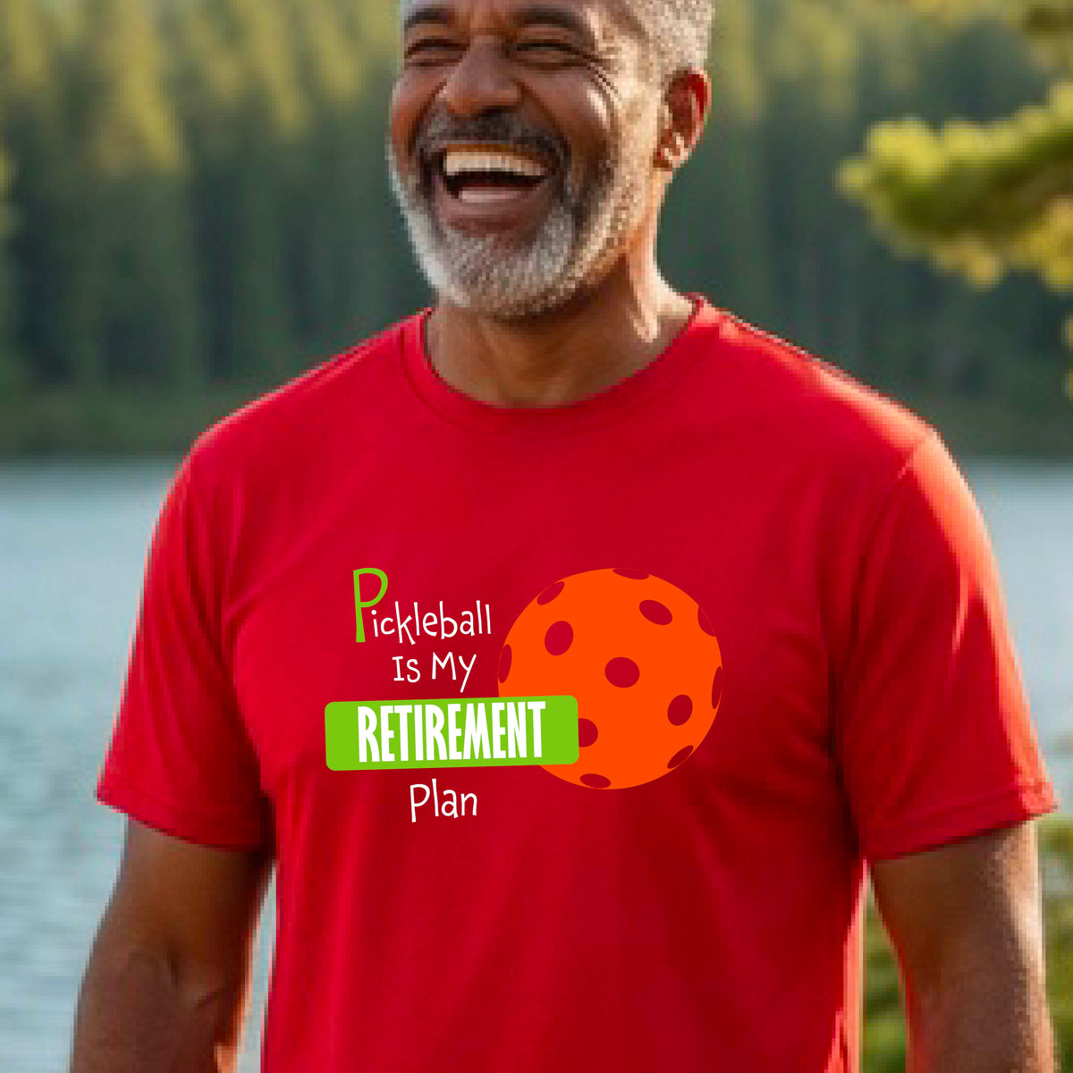 A man on a pickleball court, wearing a red short-sleeve athletic t-shirt, with the words “Pickleball is my Retirement Plan” in green and white with an orange pickleball illustration in the background
