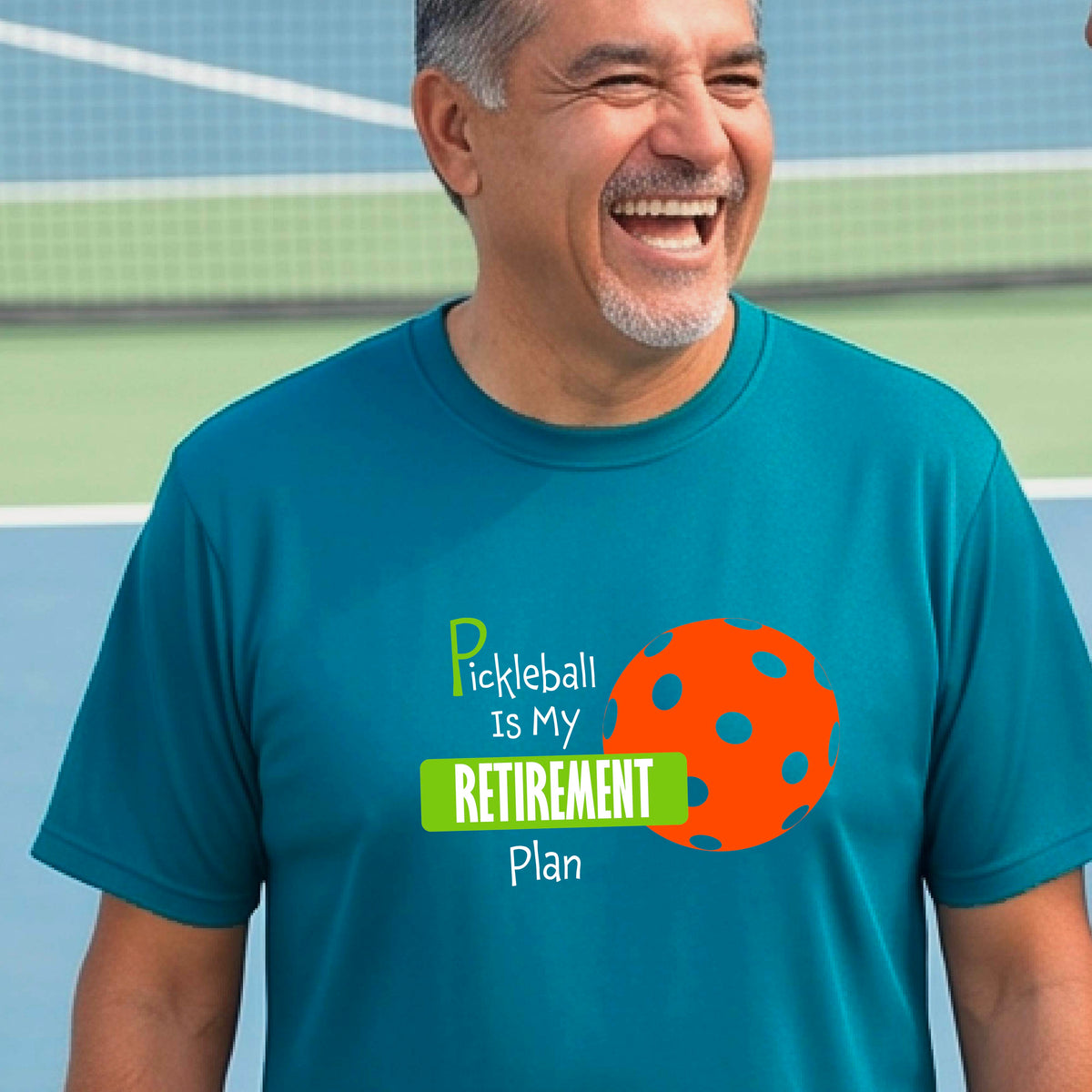 A man on a pickleball court, wearing a teal short-sleeve athletic t-shirt, with the words “Pickleball is my Retirement Plan” in green and white with an orange pickleball illustration in the background