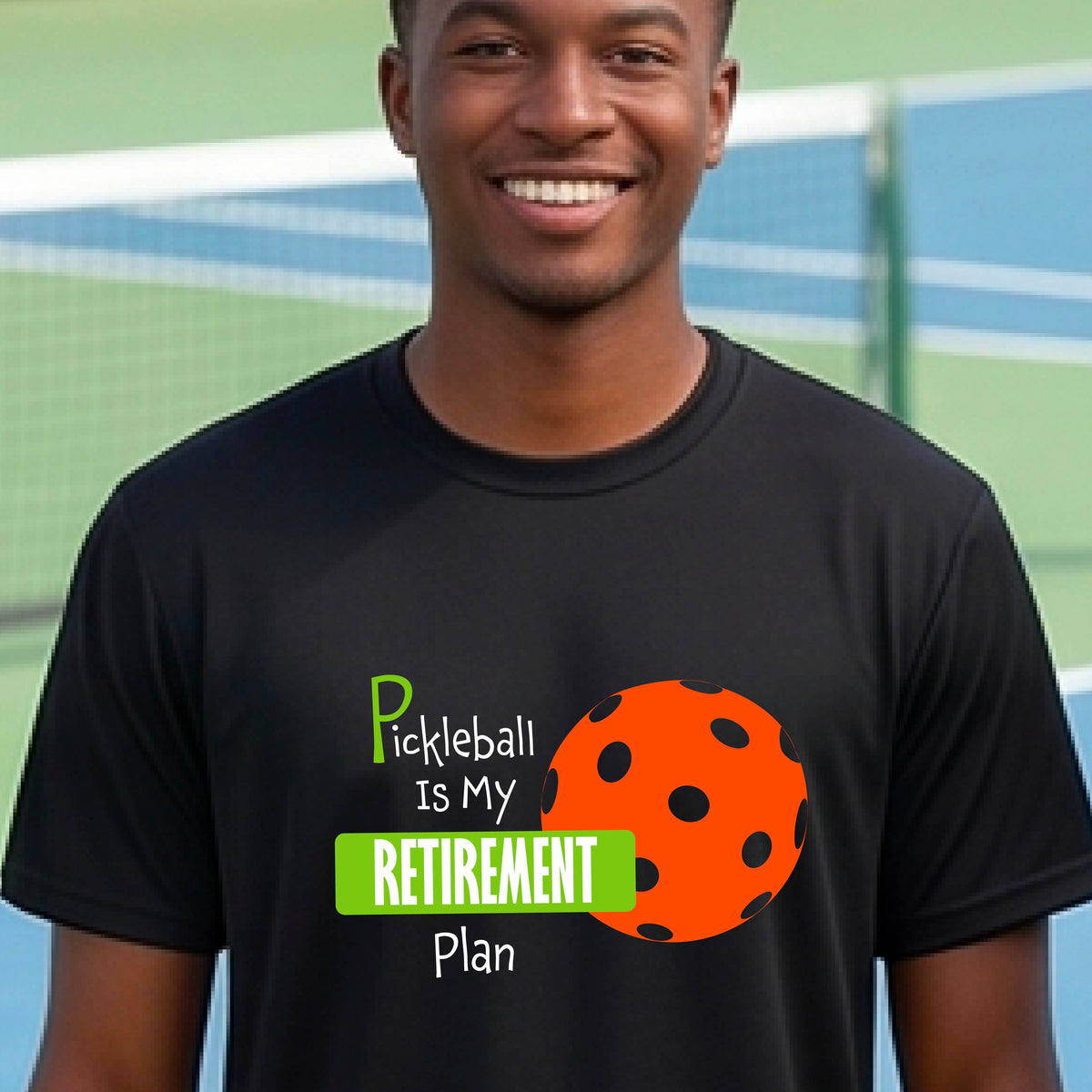A man on a pickleball court, wearing a black short-sleeve athletic t-shirt, with the words “Pickleball is my Retirement Plan” in green and white with an orange pickleball illustration in the background