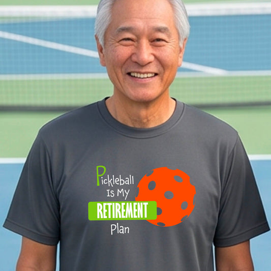 A man on a pickleball court, wearing a dark gray short-sleeve athletic t-shirt, with the words “Pickleball is my Retirement Plan” in green and white with an orange pickleball illustration in the background