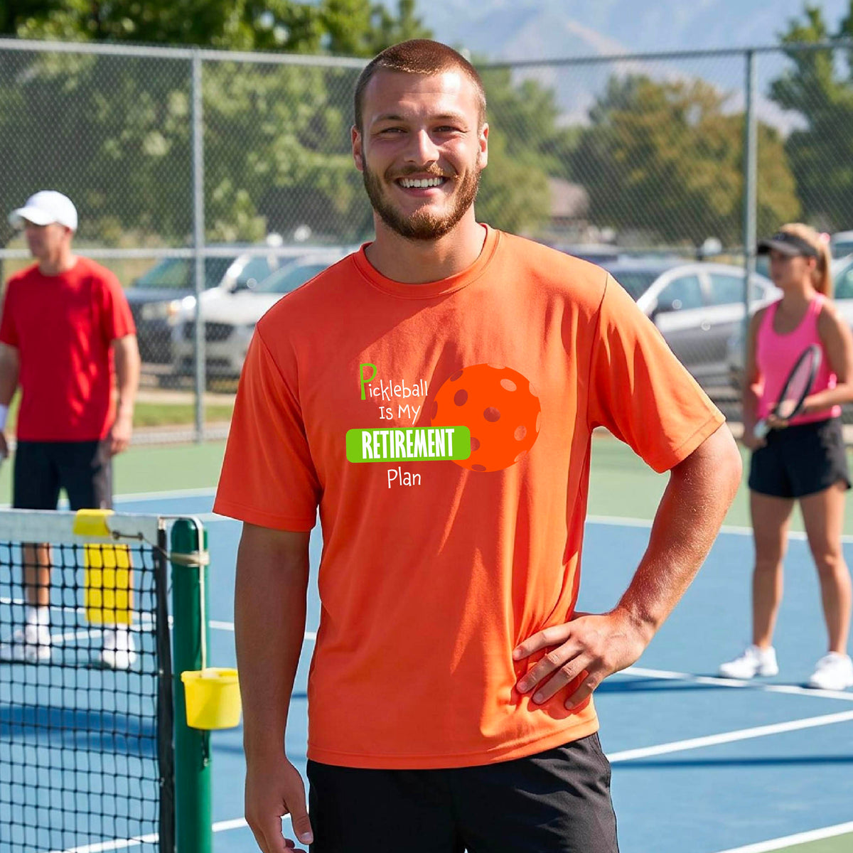 A man on a pickleball court, wearing a bright orange short-sleeve athletic t-shirt, with the words “Pickleball is my Retirement Plan” in green and white with an orange pickleball illustration in the background