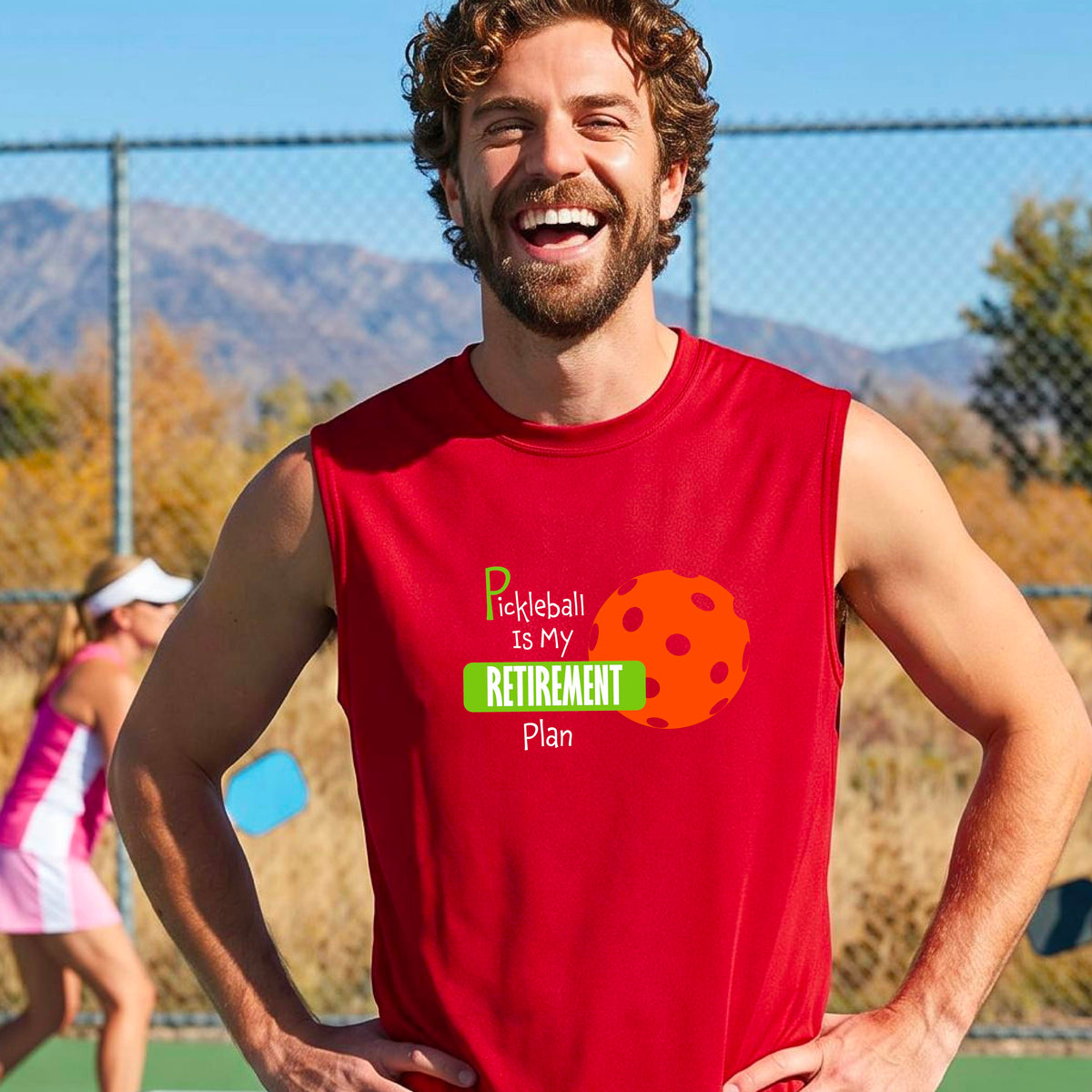 A man on a pickleball court, wearing a red sleeveless athletic t-shirt, with the words “Pickleball is my Retirement Plan” in green and white with an orange pickleball illustration in the background.