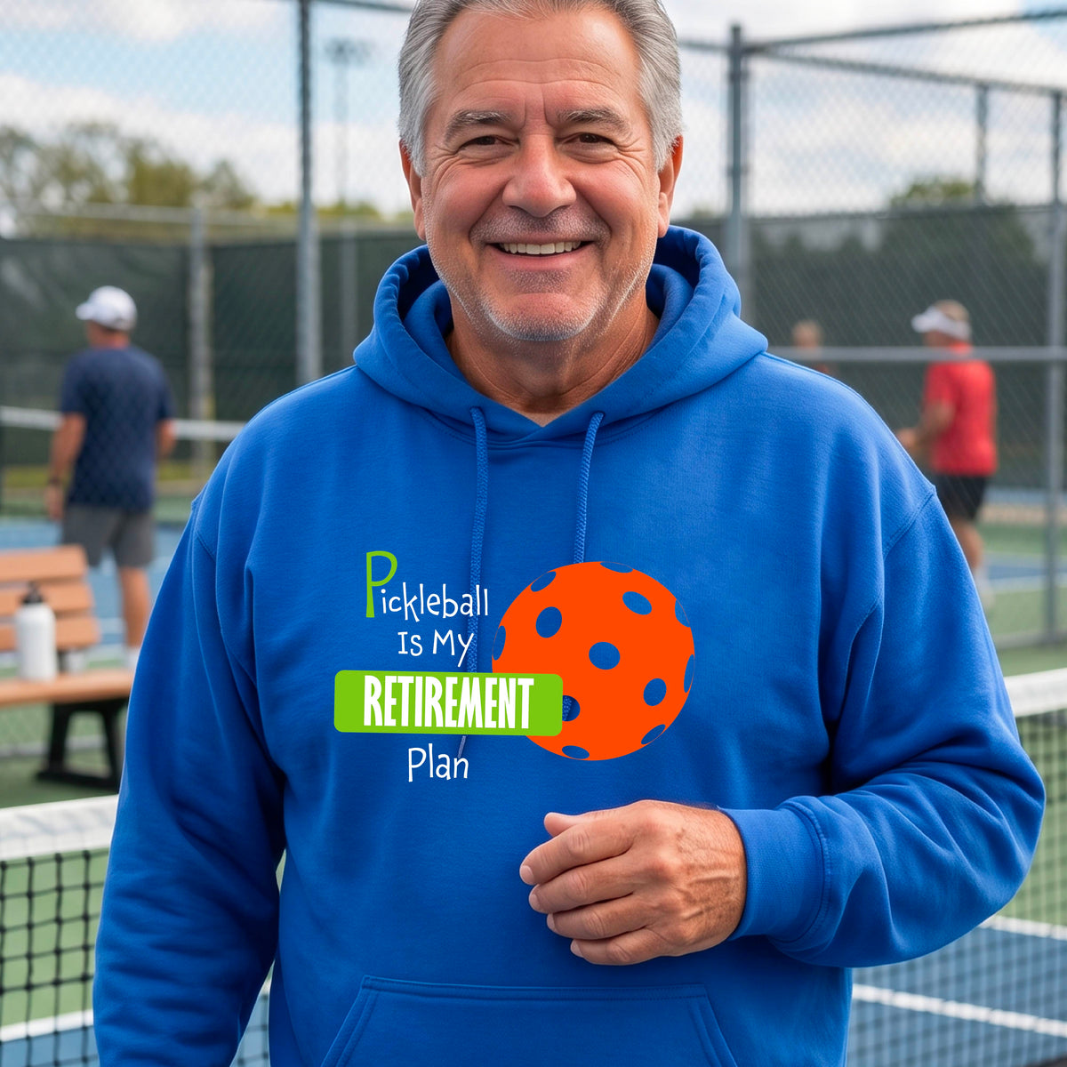 A smiling man wearing a royal blue hoodie with a graphic that reads 'Pickleball is my Retirement Plan' alongside an orange pickleball illustration, set against a blurred outdoor pickleball court background.