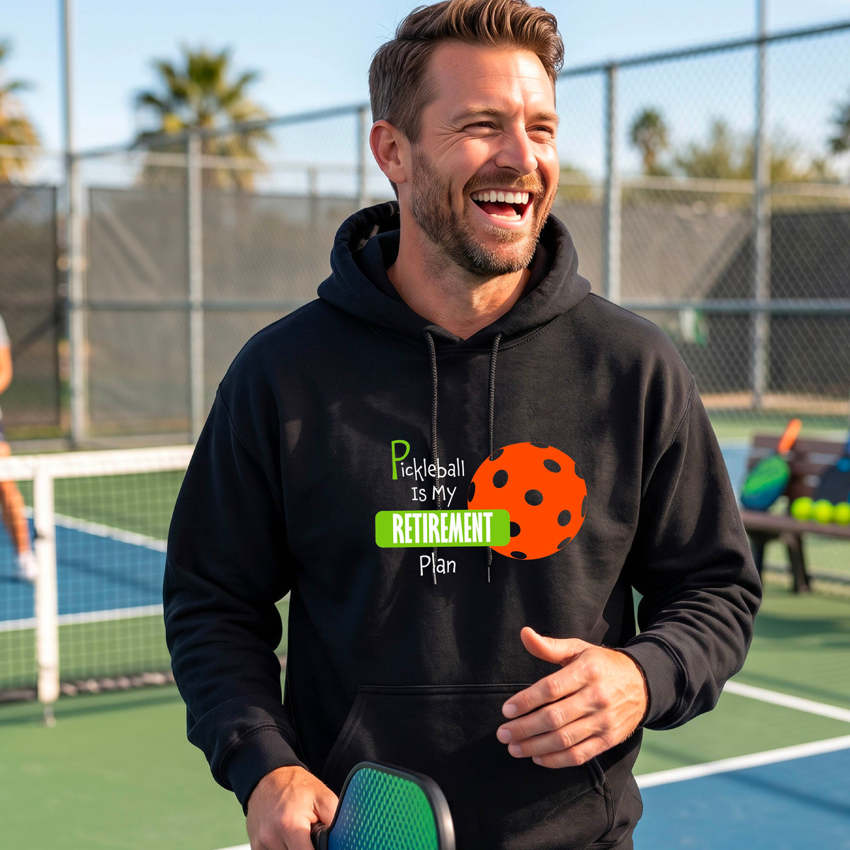 A smiling man wearing a black hoodie with a graphic that reads 'Pickleball is my Retirement Plan' alongside an orange pickleball illustration, set against a blurred outdoor pickleball court background.