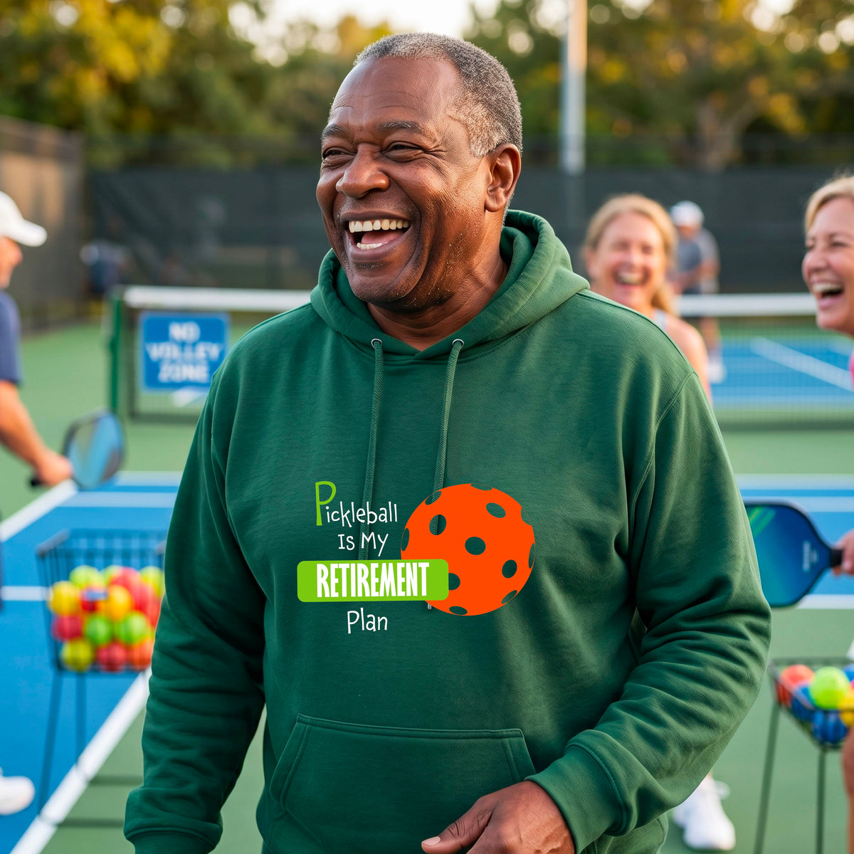 A smiling man wearing a green hoodie with a graphic that reads 'Pickleball is my Retirement Plan' alongside an orange pickleball illustration, set against a blurred outdoor pickleball court background.