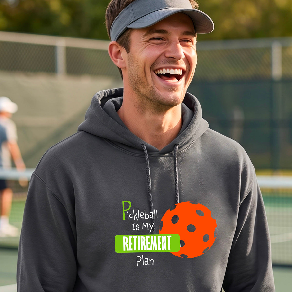 A smiling man wearing a dark gray hoodie with a graphic that reads 'Pickleball is my Retirement Plan' alongside an orange pickleball illustration, set against a blurred outdoor pickleball court background.
