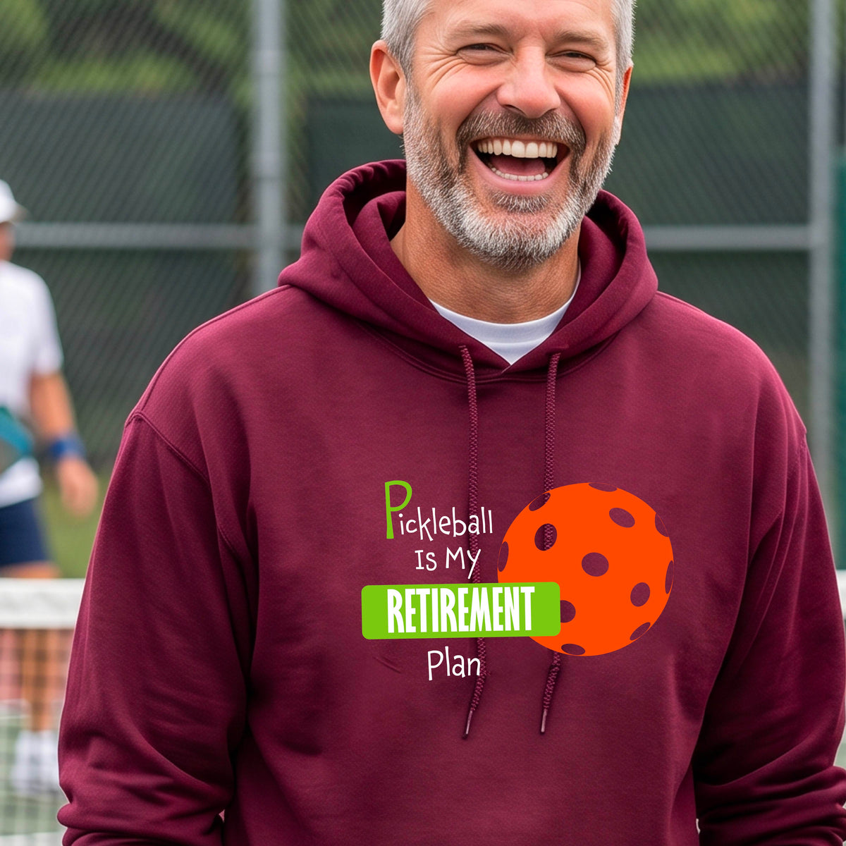 A smiling man wearing a maroon hoodie with a graphic that reads 'Pickleball is my Retirement Plan' alongside an orange pickleball illustration, set against a blurred outdoor pickleball court background.