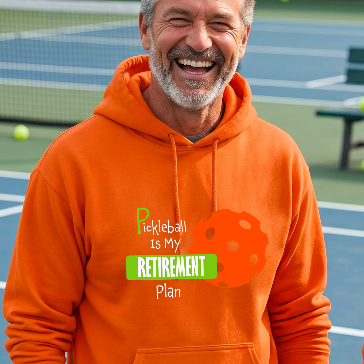 A smiling man wearing a bright orange hoodie with a graphic that reads 'Pickleball is my Retirement Plan' alongside an orange pickleball illustration, set against a blurred outdoor pickleball court background.