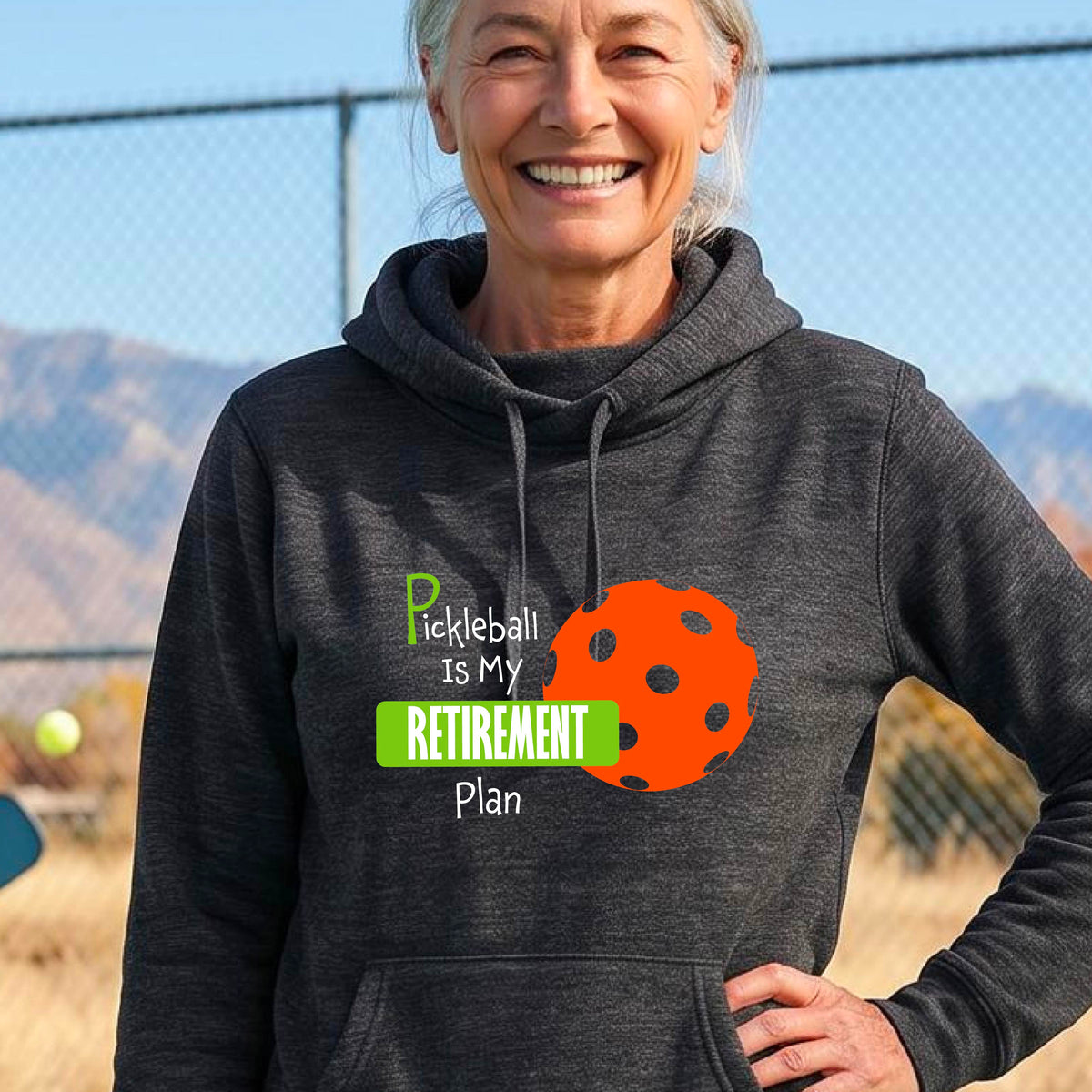 A woman on a pickleball court, wearing a black cowl-neck hoodie pullover, with the words “Pickleball is my Retirement Plan” in green and white with an orange pickleball illustration in the background