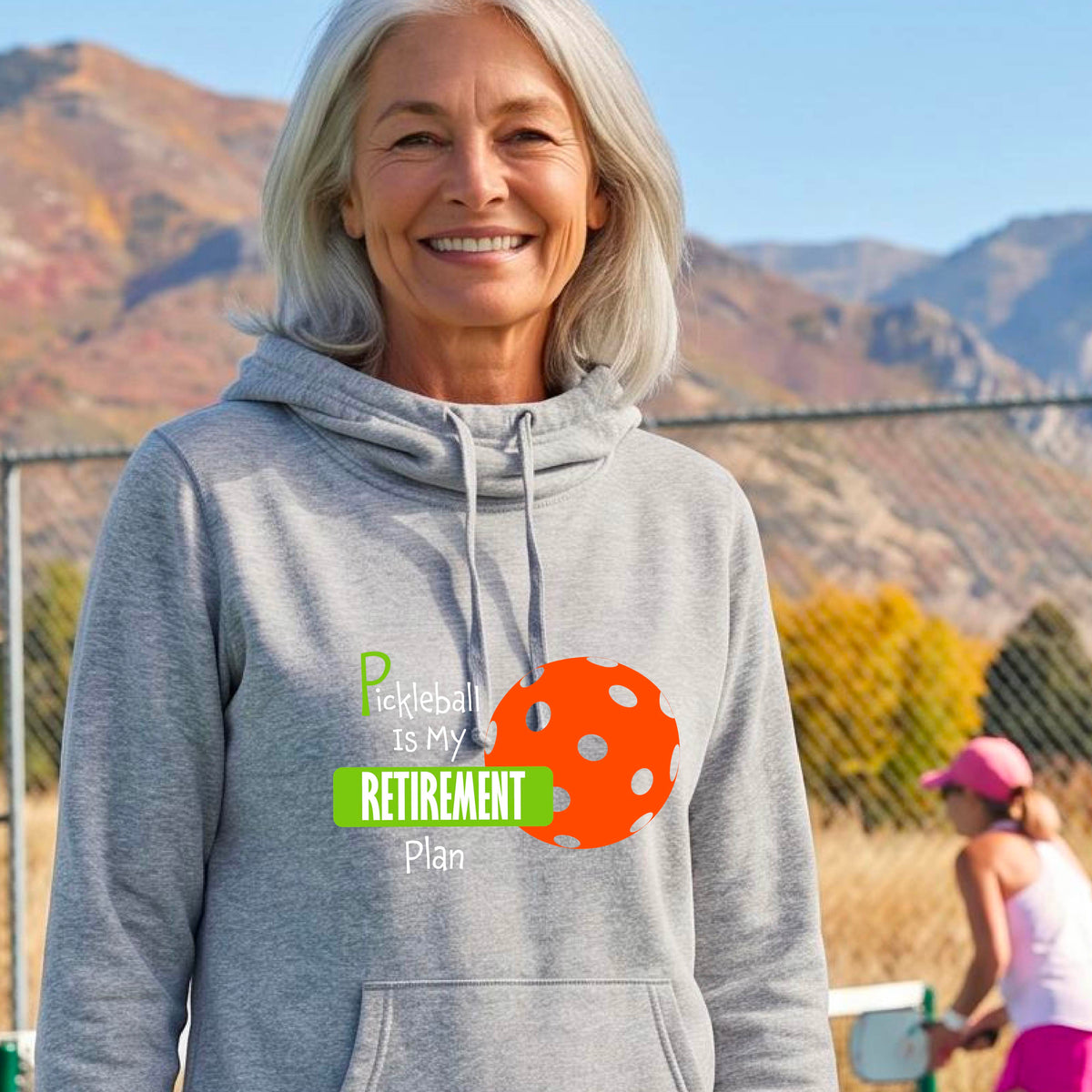 A woman on a pickleball court, wearing a light gray cowl-neck hoodie pullover, with the words “Pickleball is my Retirement Plan” in green and white with an orange pickleball illustration in the background