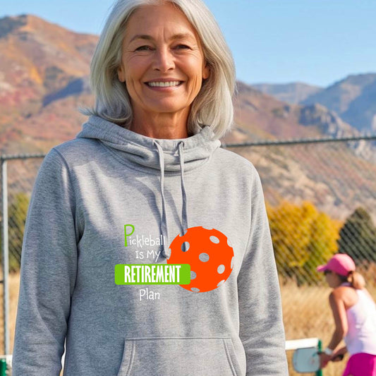 A woman on a pickleball court, wearing a light gray cowl-neck hoodie pullover, with the words “Pickleball is my Retirement Plan” in green and white with an orange pickleball illustration in the background