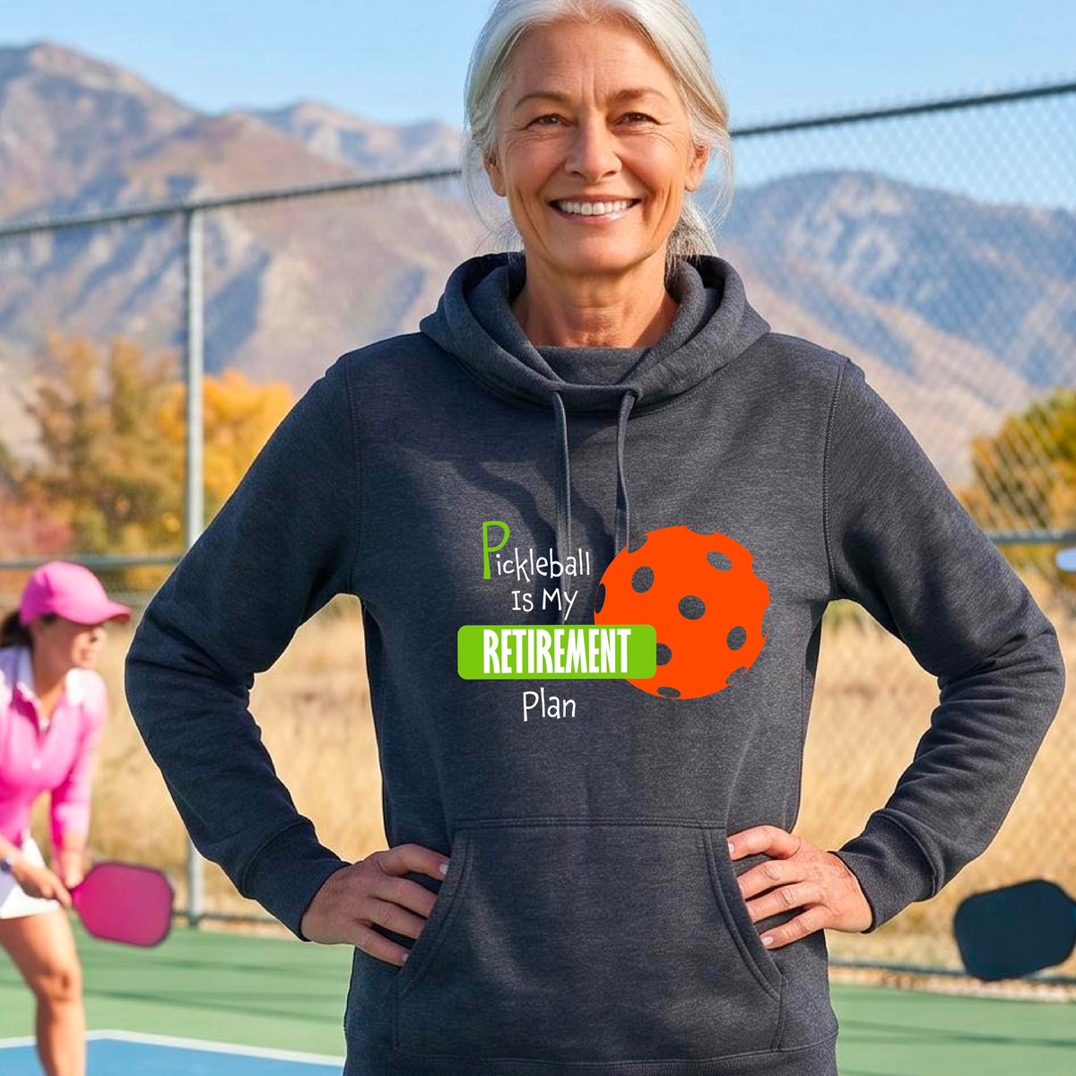 A woman on a pickleball court, wearing a navy blue heather cowl-neck hoodie pullover, with the words “Pickleball is my Retirement Plan” in green and white with an orange pickleball illustration in the background