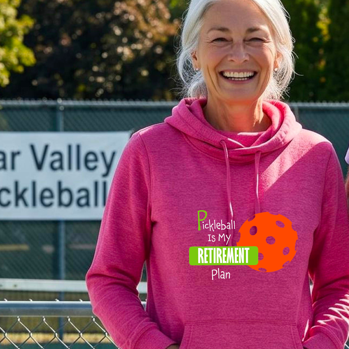 A woman on a pickleball court, wearing a pink cowl-neck hoodie pullover, with the words “Pickleball is my Retirement Plan” in green and white with an orange pickleball illustration in the background