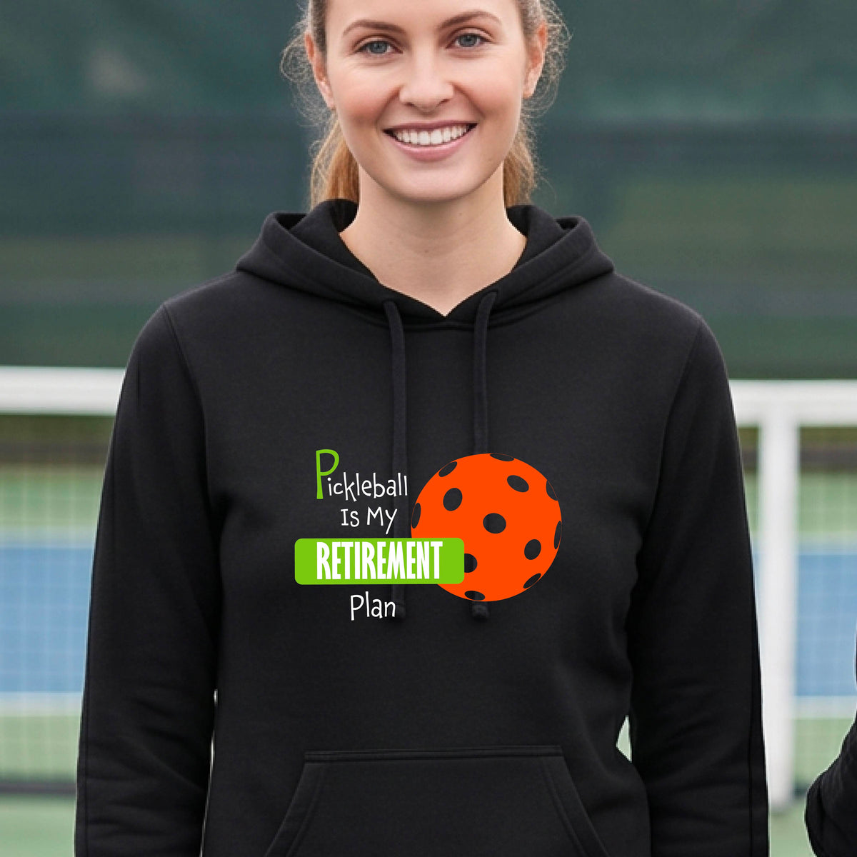 A woman smiling on a pickleball court wearing a black hoodie with a graphic that reads 'Pickleball Is My Retirement Plan' alongside an orange pickleball illustration.